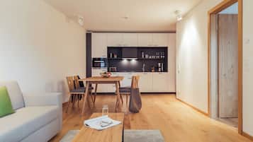 A modern living room with a sofa, a wooden coffee table, and a dining table with chairs. A kitchen with white cabinets and a black countertop is visible in the background.