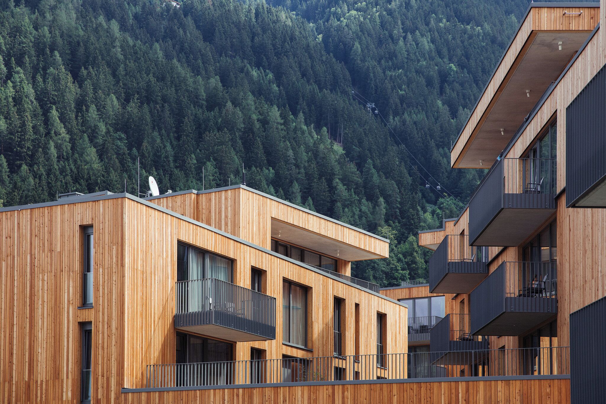 A modern multi-story building with wooden paneling, balconies, and a forested hillside in the background.