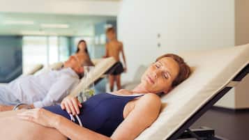 A person lying on a sunbed in a room with a glass wall, another person walking in the background, and a third person standing near the glass wall.