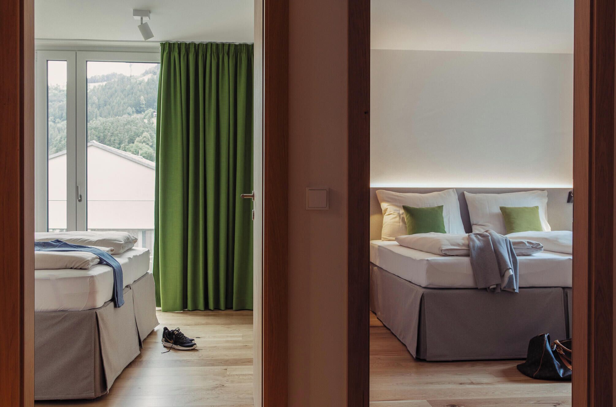 Habitación de hotel con cama, ventana con cortinas, piso de madera y vistas a zona boscosa.