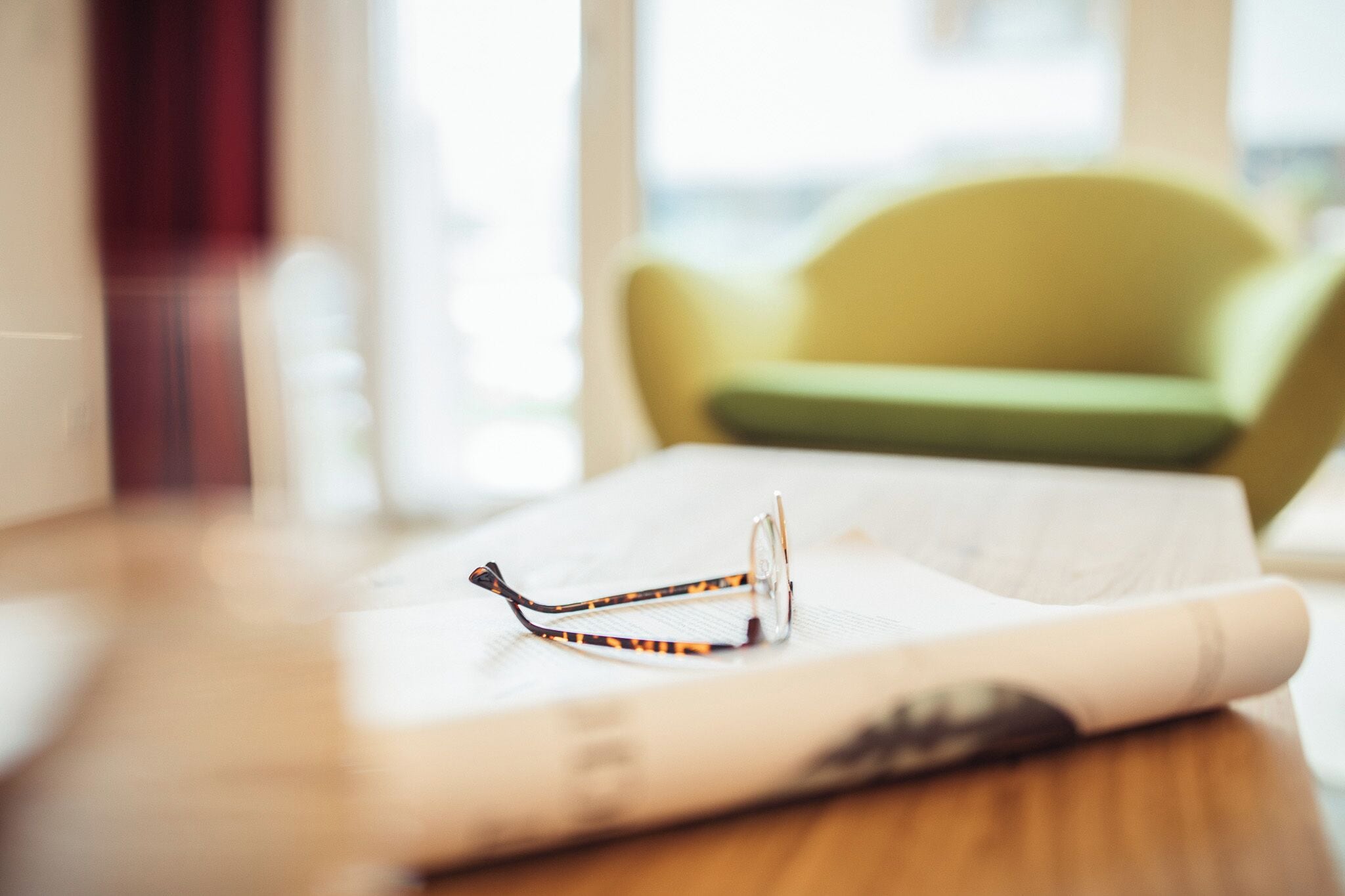 A pair of glasses on a white surface, possibly a table, with a blurred background featuring a green chair.