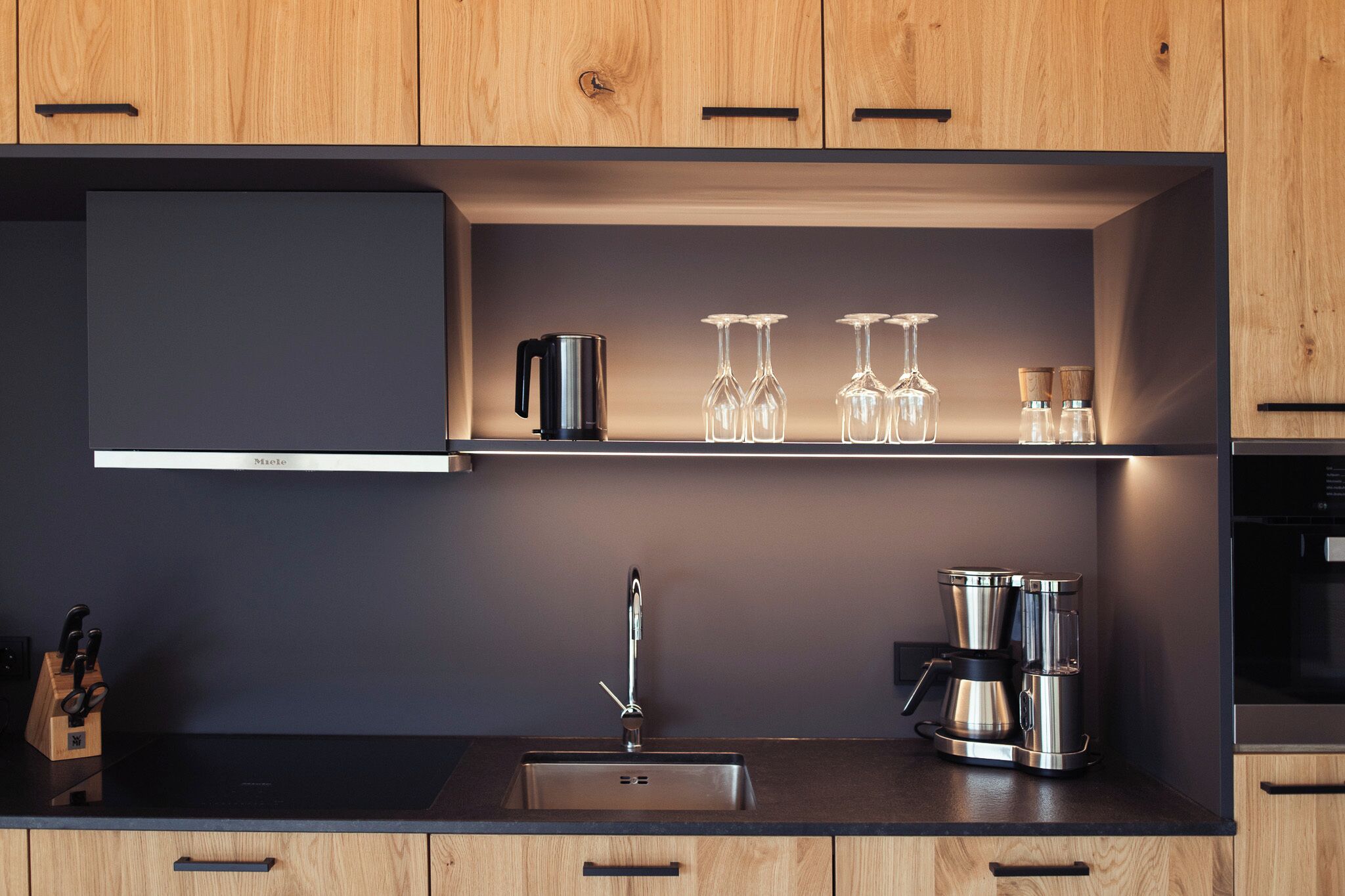 Una cocina moderna con armarios de madera, encimera negra, horno empotrado, cafetera y copas de vino en un estante.