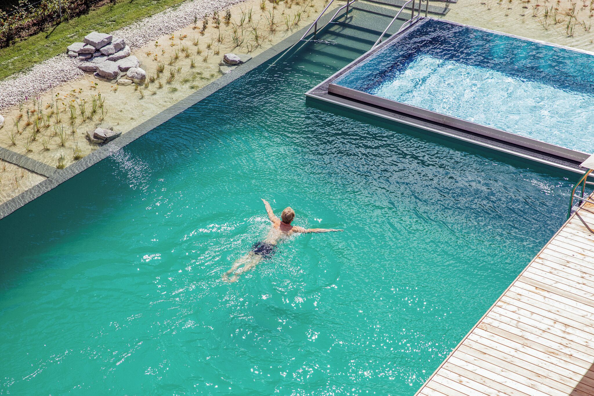 A person swimming in a clear blue pool with a wooden deck and a small garden area with rocks and plants.