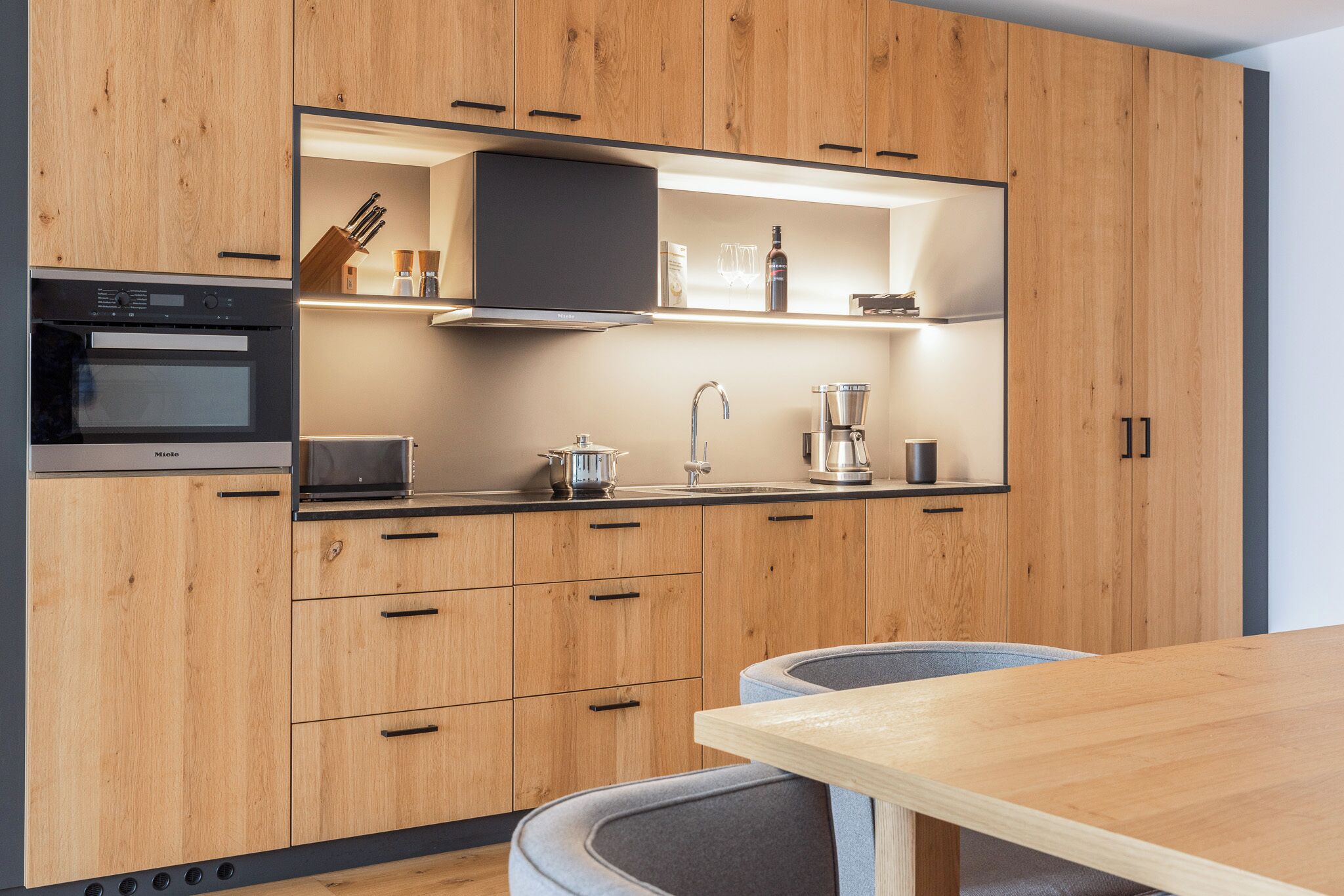 Una cocina moderna con armarios de madera, horno empotrado, microondas, cafetera y un comedor con mesa y sillas.