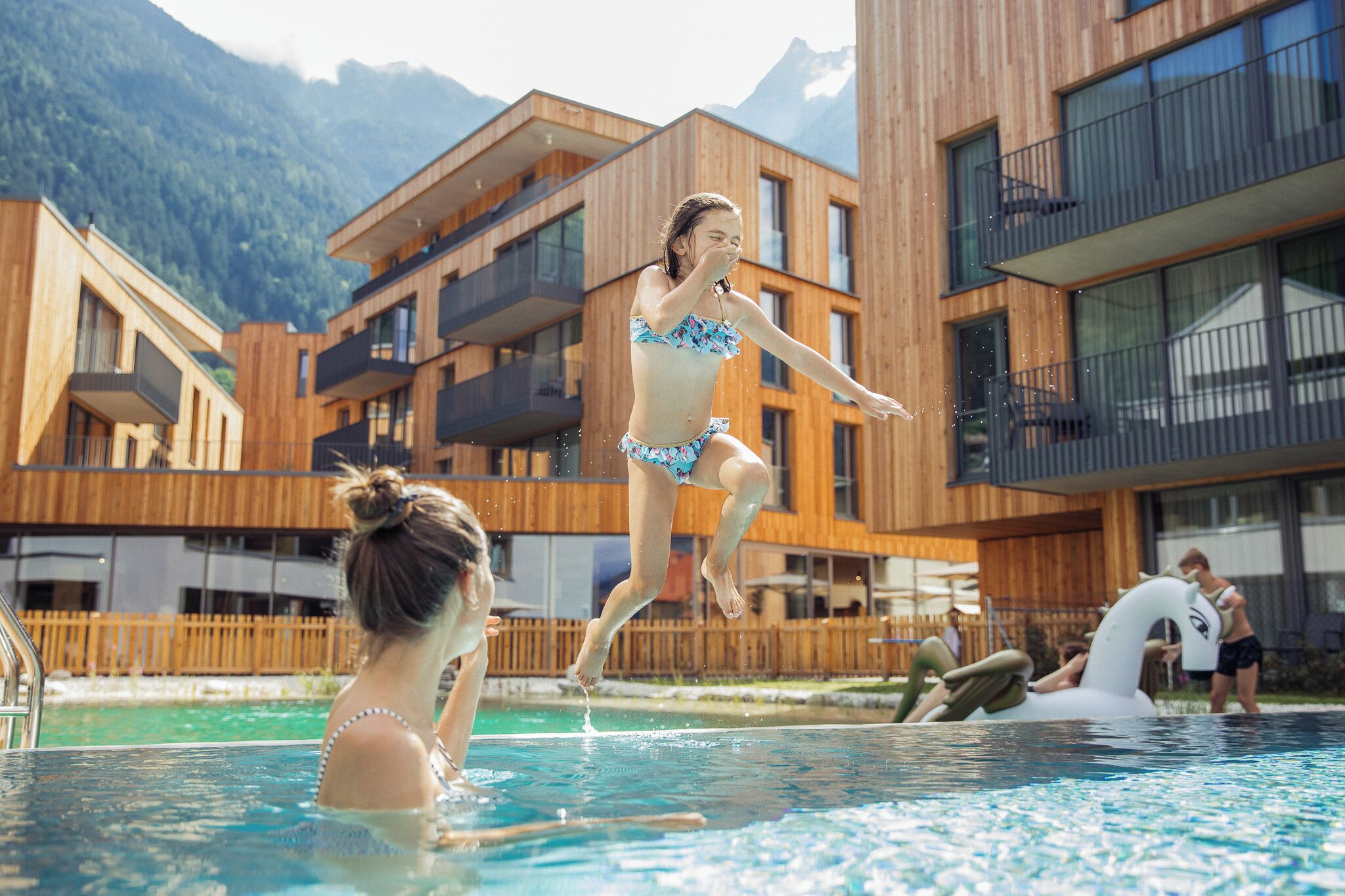 Una piscina con gente disfrutando del agua, rodeada de edificios modernos de madera con balcones.