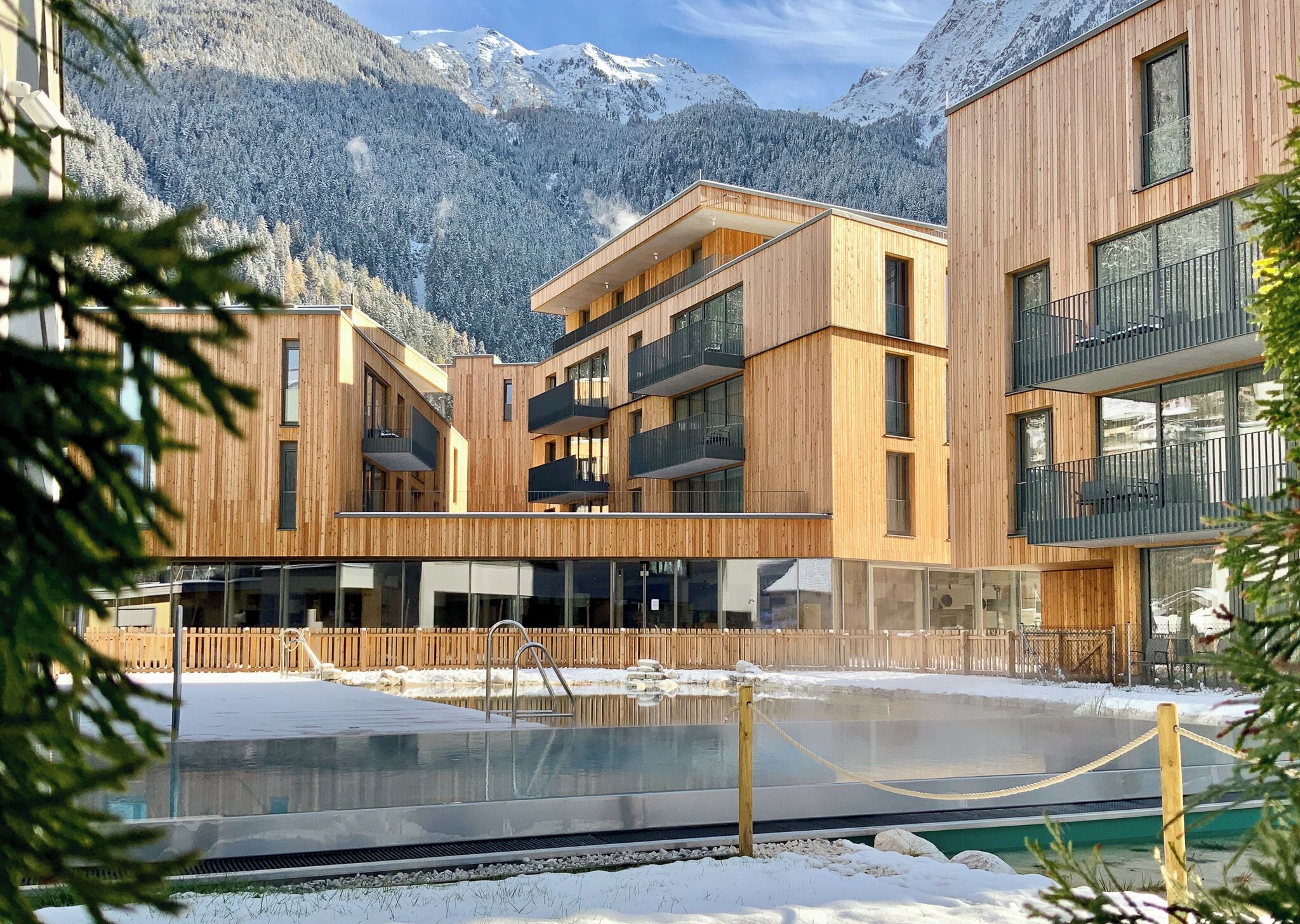Un edificio moderno con paredes exteriores de madera, piscina y vistas a montañas nevadas.