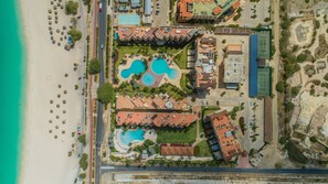 An aerial view of a resort with multiple swimming pools, a beach, and a parking area.