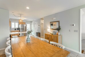A spacious dining area with a wooden table and white chairs, a kitchen in the background, and a light-colored wall with a mirror and decorative items.