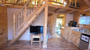 A wooden interior with a TV on a small stand, a kitchen area with a sink and appliances, and a staircase with exposed beams. - Sunset Ridge #2 at Rustic Ridge (Keystone)
