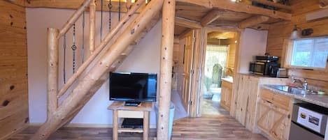 A wooden interior with a TV on a small stand, a kitchen area with a sink and appliances, and a staircase with exposed beams.