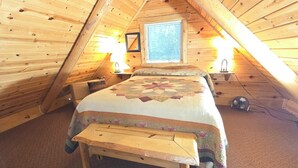 A cozy attic bedroom with a wooden sloped ceiling, a large bed with a floral quilt, two wall-mounted lamps, and a window with blinds. - Sunset Ridge #2 at Rustic Ridge (Keystone)