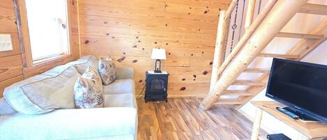 A cozy cabin interior with a wooden floor, a sofa with patterned cushions, a small black heater, a wooden TV stand, and a wooden staircase.