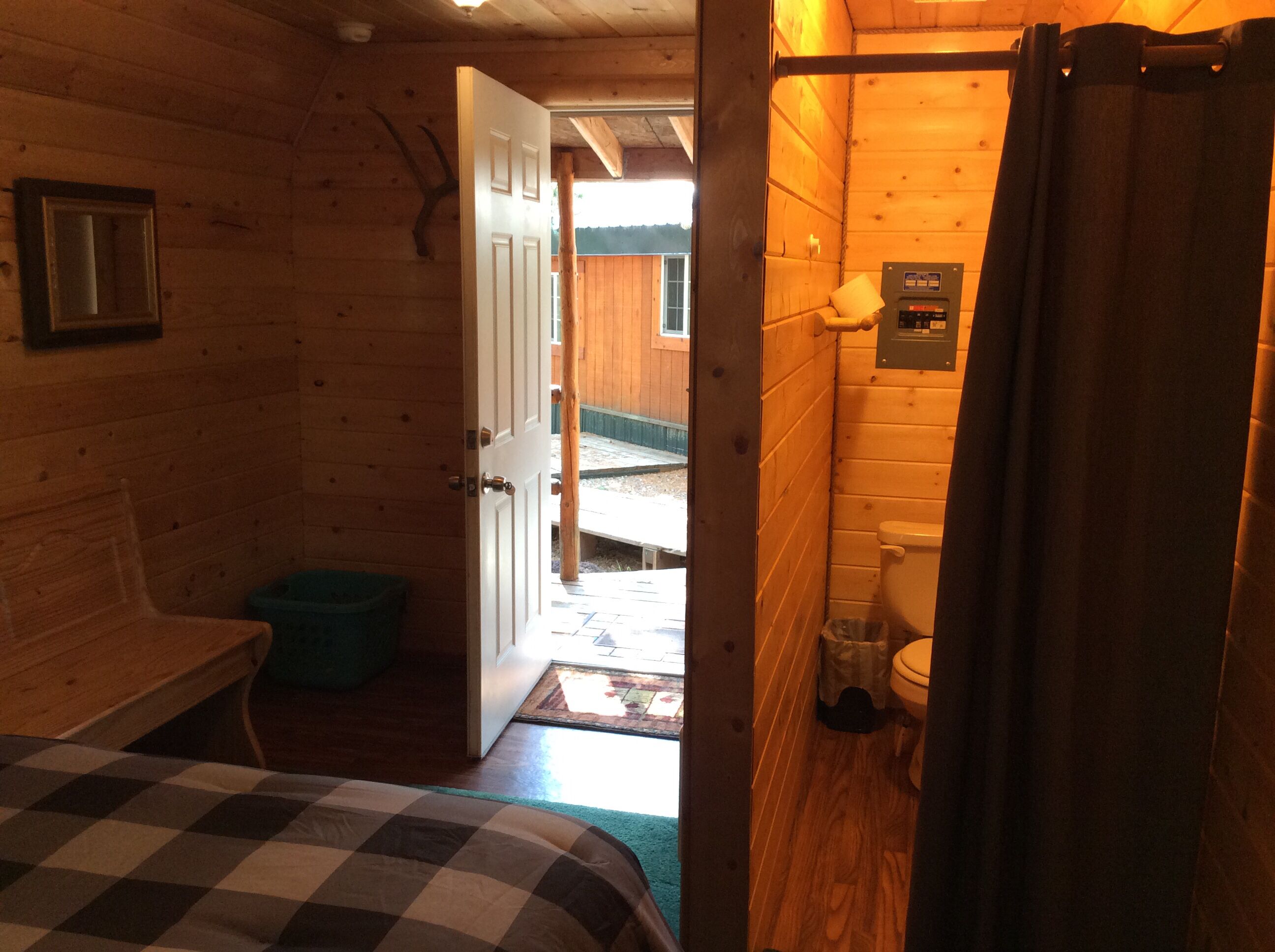 A wooden cabin interior with a bed, a small table, and a bathroom with a toilet and shower curtain.