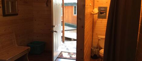 A wooden cabin interior with a bed, a small table, and a bathroom with a toilet and shower curtain.