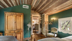 Une pièce avec un plafond en bois, une grande armoire, un lit recouvert d’une couvre-lit à motifs, une chaise et une fenêtre donnant sur des arbres.