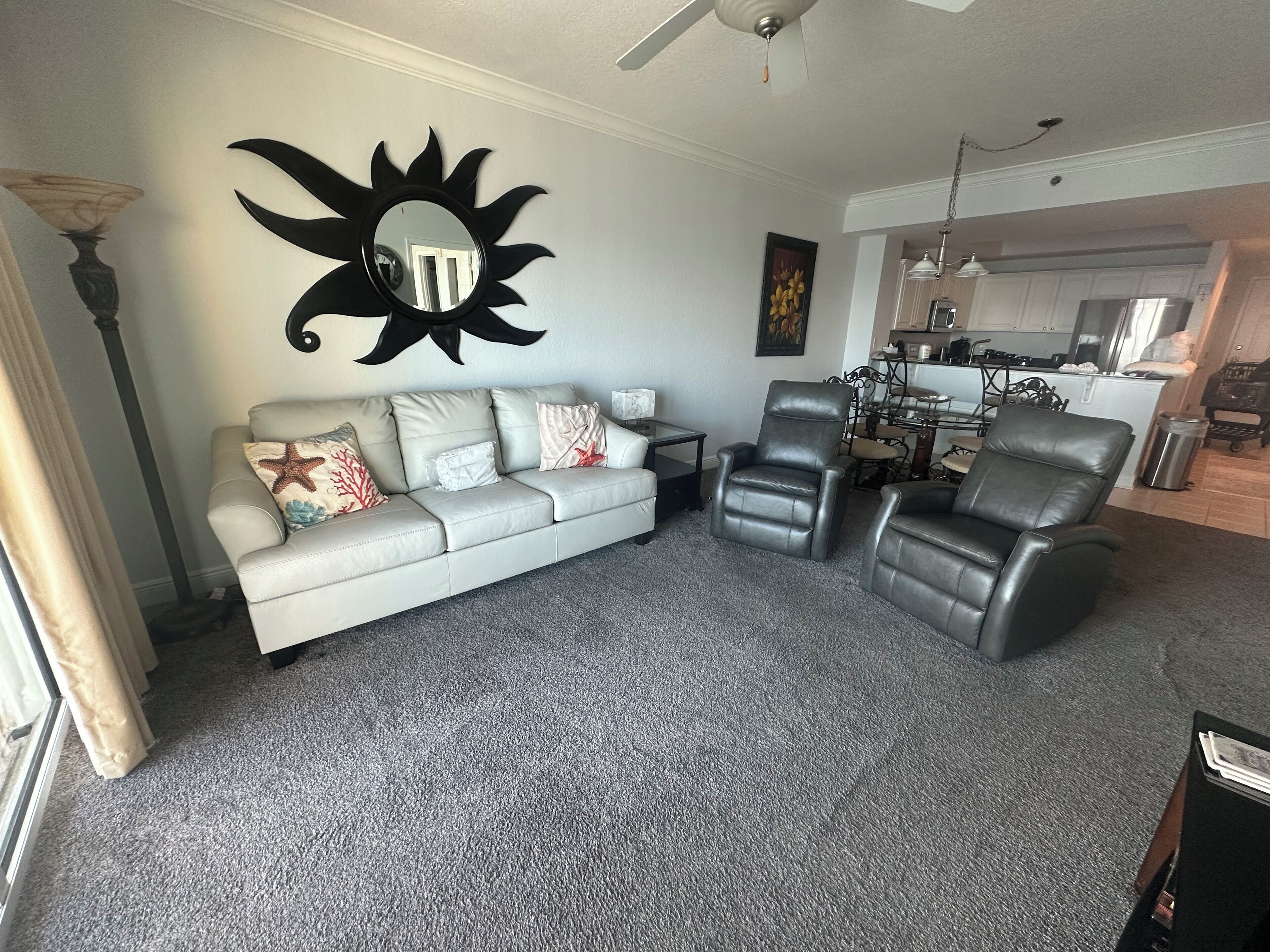 A living room with a grey sofa, two black leather chairs, a sun-shaped mirror, and a kitchen in the background.