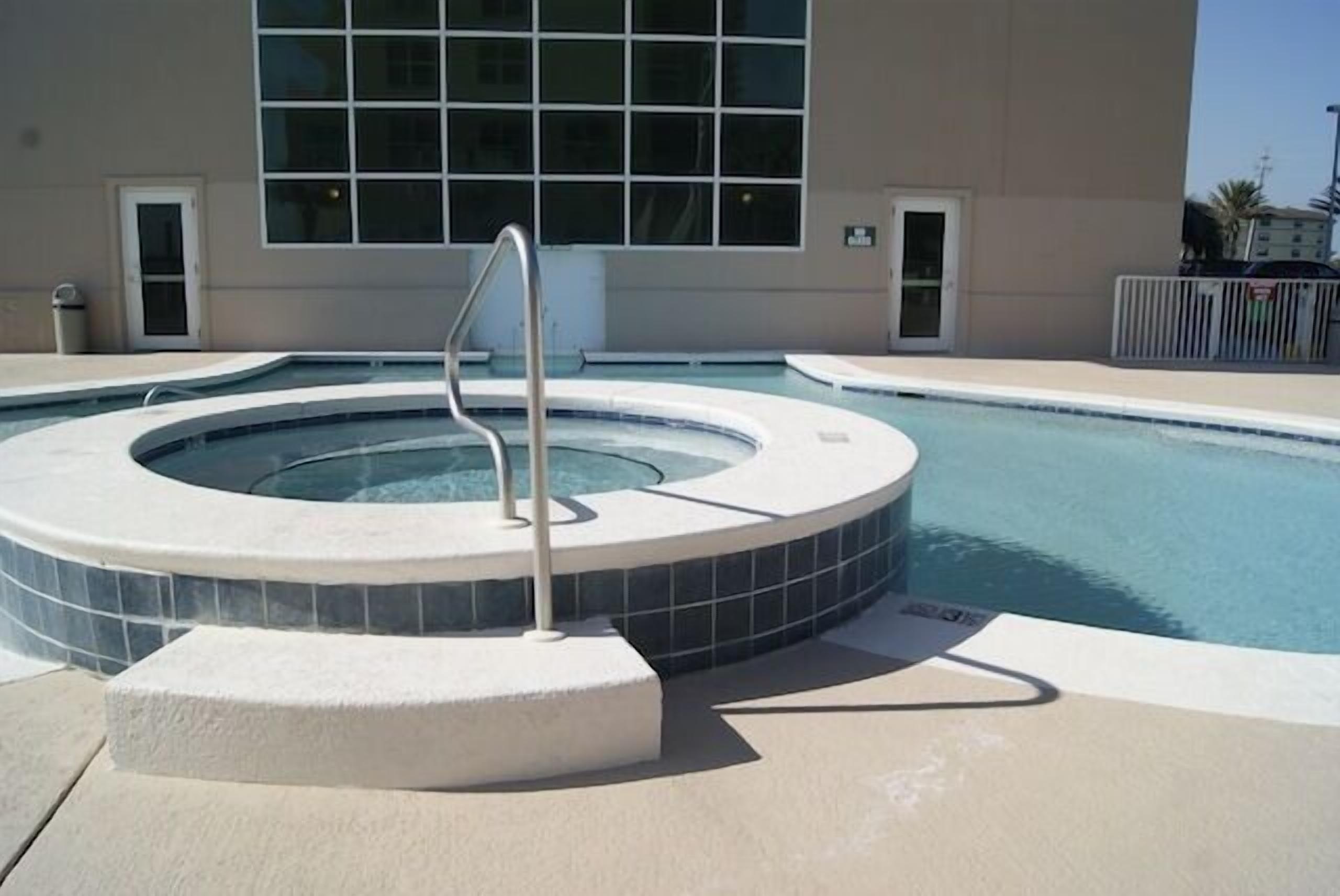 A swimming pool with a hot tub, surrounded by a beige deck, and a building with large windows in the background.