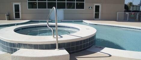 A swimming pool with a hot tub, surrounded by a beige deck, and a building with large windows in the background.