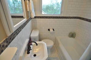 Ein Badezimmer mit weißem Waschbecken, Toilette und Badewanne, einem Fenster mit Blick auf Bäume und einem gefliesten Boden.