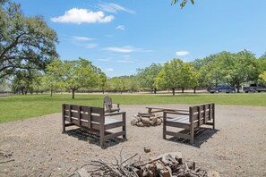 An outdoor seating area with two wooden benches, a fire pit, and a clear view of a park with trees and vehicles in the background.