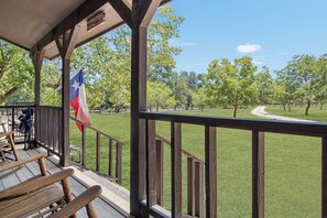 A covered outdoor area with a wooden deck, a flag, and a view of a grassy field with trees. - Double U Barr Texan | Cabin with Creek Access (Bandera)