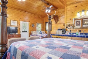 A cozy cabin interior with a large bed, a television, a wooden ceiling, and a mounted animal head. - Double U Barr Texan | Cabin with Creek Access (Bandera)