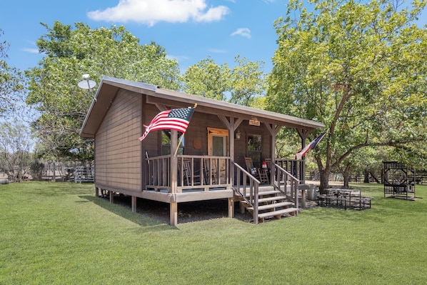 A small wooden cabin with a porch and an American flag. - Double U Barr Texan | Cabin with Creek Access (Bandera)