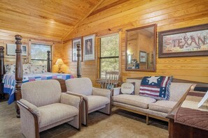 A cozy cabin interior with a wooden ceiling, a large framed 'TEXAN' sign, a bed with a quilt, and a sofa with a 'TEXAS' pillow.