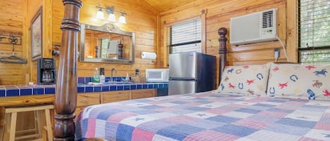 A cozy cabin-style room with a wooden ceiling, a bed with a quilt, a wooden chair, a small table, a mirror, a refrigerator, a microwave, and a window with blinds.