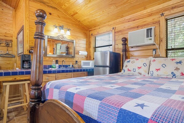A cozy cabin-style room with a wooden ceiling, a bed with a quilt, a wooden chair, a small table, a mirror, a refrigerator, a microwave, and a window with blinds.