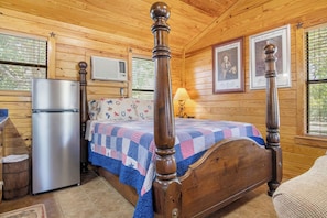 A wooden cabin bedroom with a bed, a refrigerator, and a window with blinds. - Double U Barr Texan | Cabin with Creek Access (Bandera)