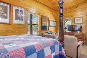 A room with a large bed, a wooden ceiling, a television, and framed pictures on the walls. - Double U Barr Texan | Cabin with Creek Access (Bandera)