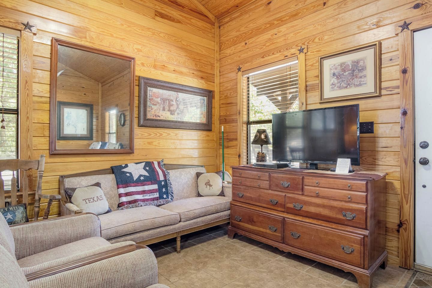 A cozy room with a wooden ceiling, a sofa with a Texas-themed pillow, a television on a wooden dresser, and framed pictures on the walls.
