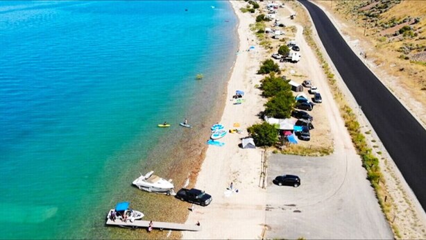 An aerial view of a beach with clear turquoise water, a paved road, and parked cars along the shore.