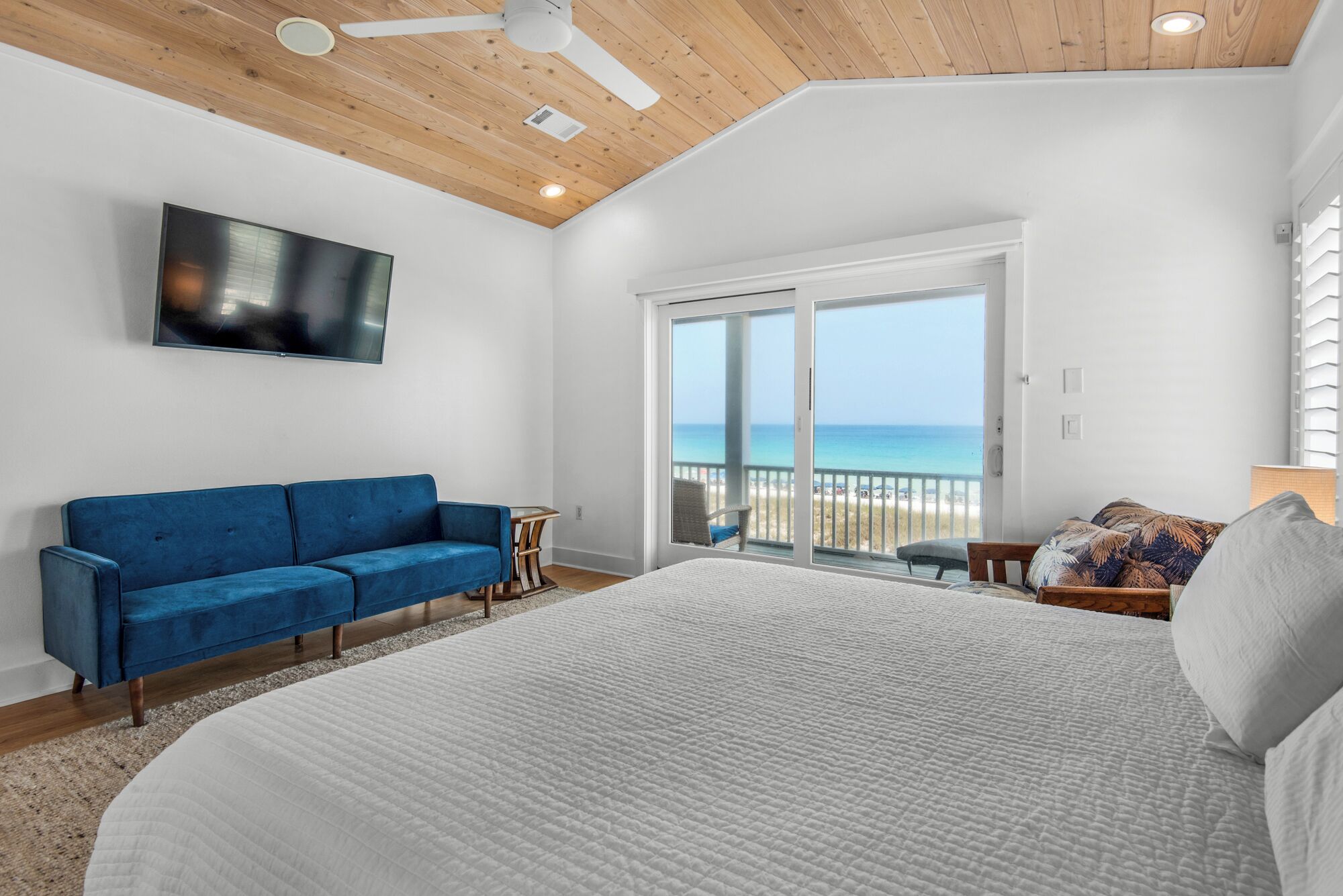 A bedroom with a large bed, a blue sofa, a TV, and a balcony with a view of the ocean.