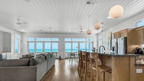 A spacious living area with a large gray sofa, wooden flooring, and a kitchen with stainless steel appliances.