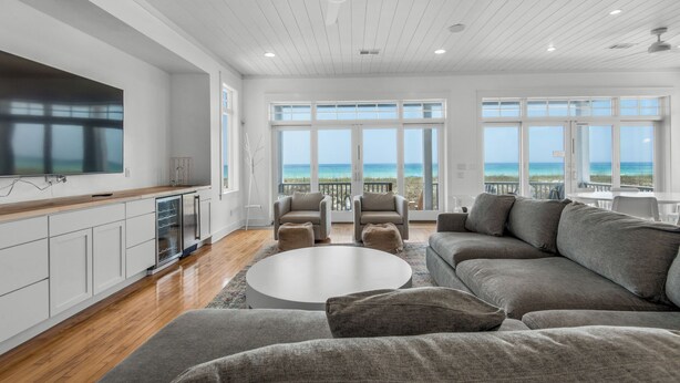 A spacious living room with a large flat-screen TV, a grey sectional sofa, and a round coffee table. There are multiple windows offering a view of the ocean.