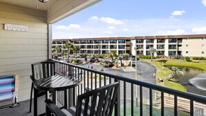 A balcony with a table and chairs, a sign reading "Blessings", and a view of a multi-story building with a landscaped area and a pond. - Ocean view, unique, stylish, contemporary, upscale condo with a modern ambiance (Hilton Head Island)