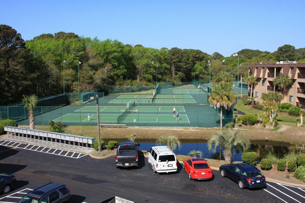 Una cancha de tenis con una valla perimetral, un área de estacionamiento con varios coches y un edificio de techo plano al fondo.
