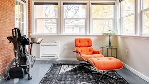 A room with a brick wall, large windows, a treadmill, an air conditioning unit, a patterned rug, and an orange chair. - Remodeled, Updated, Perfect Location Next to Downtown/PSU! (State College)