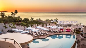 Zwembad van een resort met ligbedden en parasols, met uitzicht op de oceaan bij zonsondergang.
