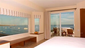 Hotelkamer met een groot raam met uitzicht op zee, een bed, een bureau en een balkon met een tafel en stoelen.