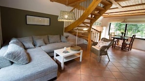 Uma sala de estar moderna com escada de madeira, sofá seccional cinza, mesa de centro branca, cadeiras de vime e um espaço de jantar com mesa e cadeiras.