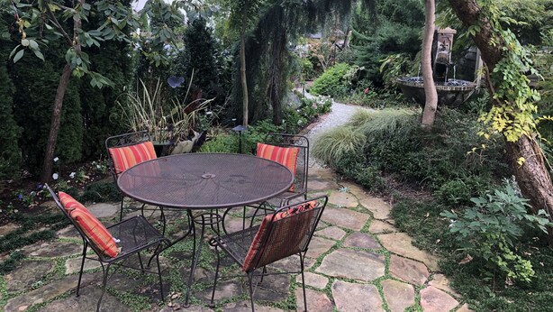 Eine Garten-Terrasse mit rundem Tisch und Stühlen, umgeben von üppiger Bepflanzung und einem Kiesweg.