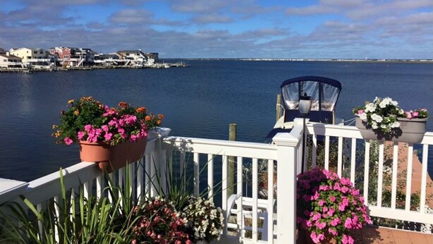 A balcony with potted flowers, a chair, and a view of a calm body of water with buildings in the distance. - LBI Bayfront Beauty Brand New Hot Tub! Boat Slip Included! Two blocks to Beach! (Ship Bottom)