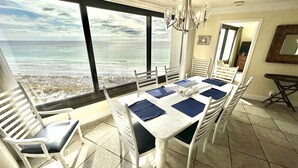Un espace repas avec vue sur l’océan, des chaises blanches, un lustre et un sol carrelé.