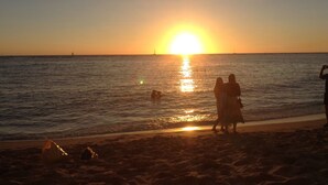 Scena di spiaggia al tramonto con il sole basso sull'orizzonte, persone che camminano lungo la riva e una barca a vela in lontananza.