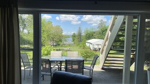 Een kamer met een groot raam dat uitkijkt op een terras met buitenmeubilair en een uitzicht op een meer en bomen.