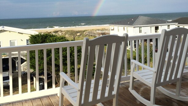 A wooden deck with white rocking chairs overlooking a beach and a rainbow. - Custom Home With Many Extras with beautiful Ocean Views! (Ocean Isle Beach)