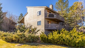 A modern two-story house with a balcony, surrounded by evergreen shrubs and a clear blue sky.